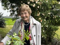 Elisabeth T. in einem Garten mit blühenden Rosensträuchern, trägt eine helle Jacke und eine Perlenkette.