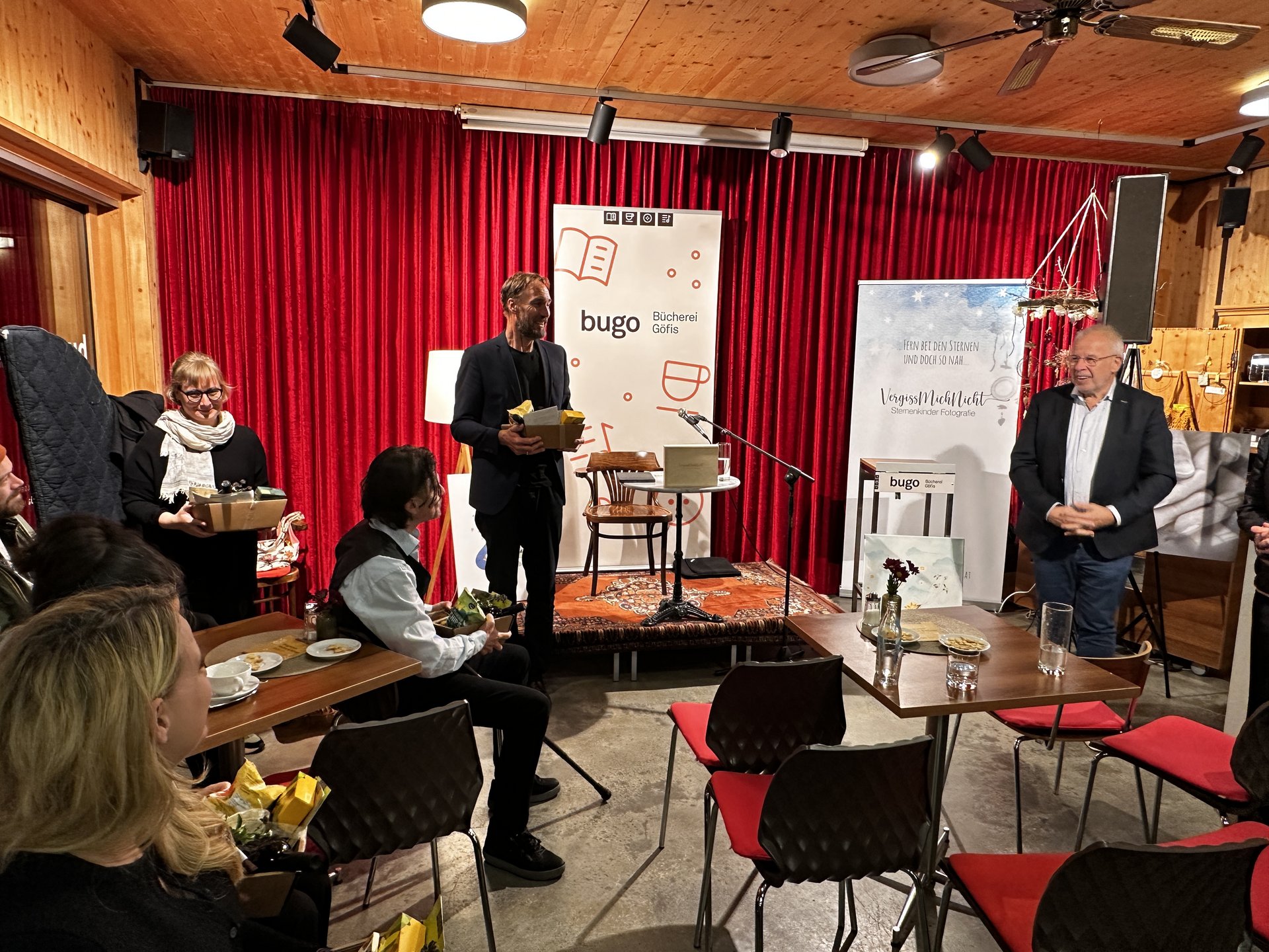 Buchlesung in der Bücherei Güfis mit Autor und Gästen vor rotem Vorhang.